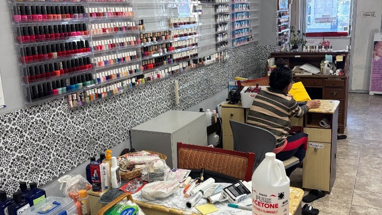 Nail service desk with technician seating; acetone and supplies visible; full wall of polish colors behind.